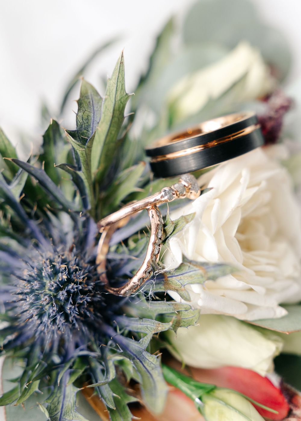 Close-up of wedding rings nestled among thistle flowers and white roses in a bridal bouquet.