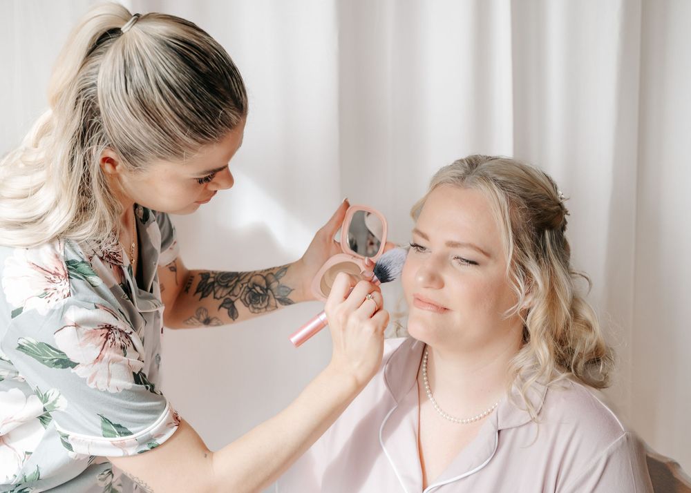 Professional makeup application being done in a bright room with white curtains.