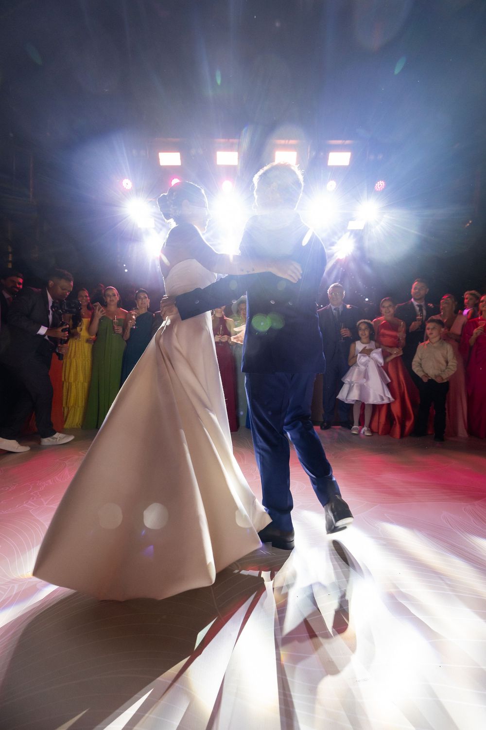 Dancing couple illuminated by bright spotlights on a ballroom dance floor during what appears to be a wedding reception.