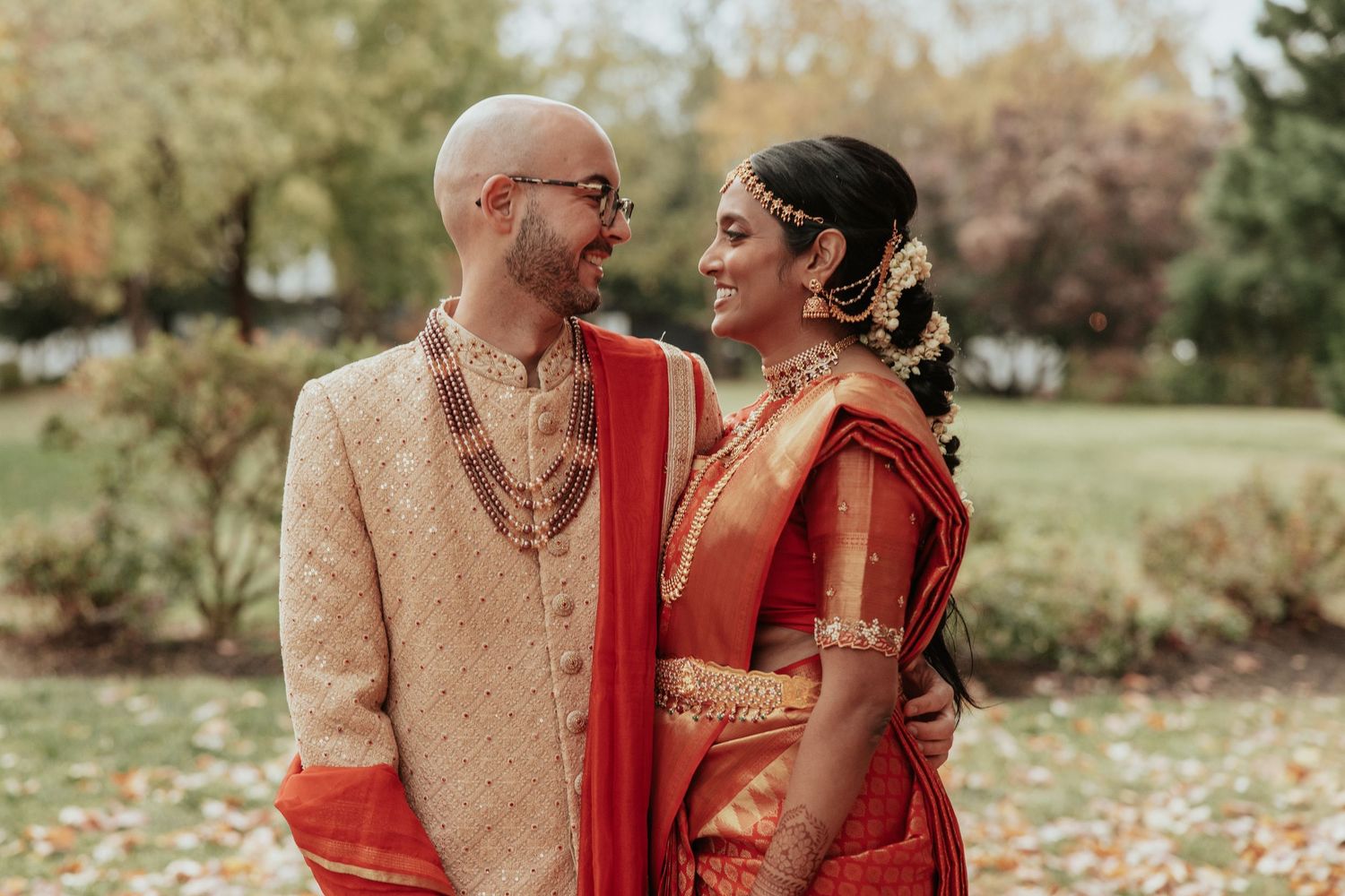 An affectionate couple in matching beige and red cultural attire share tender moments in a park with autumn foliage.