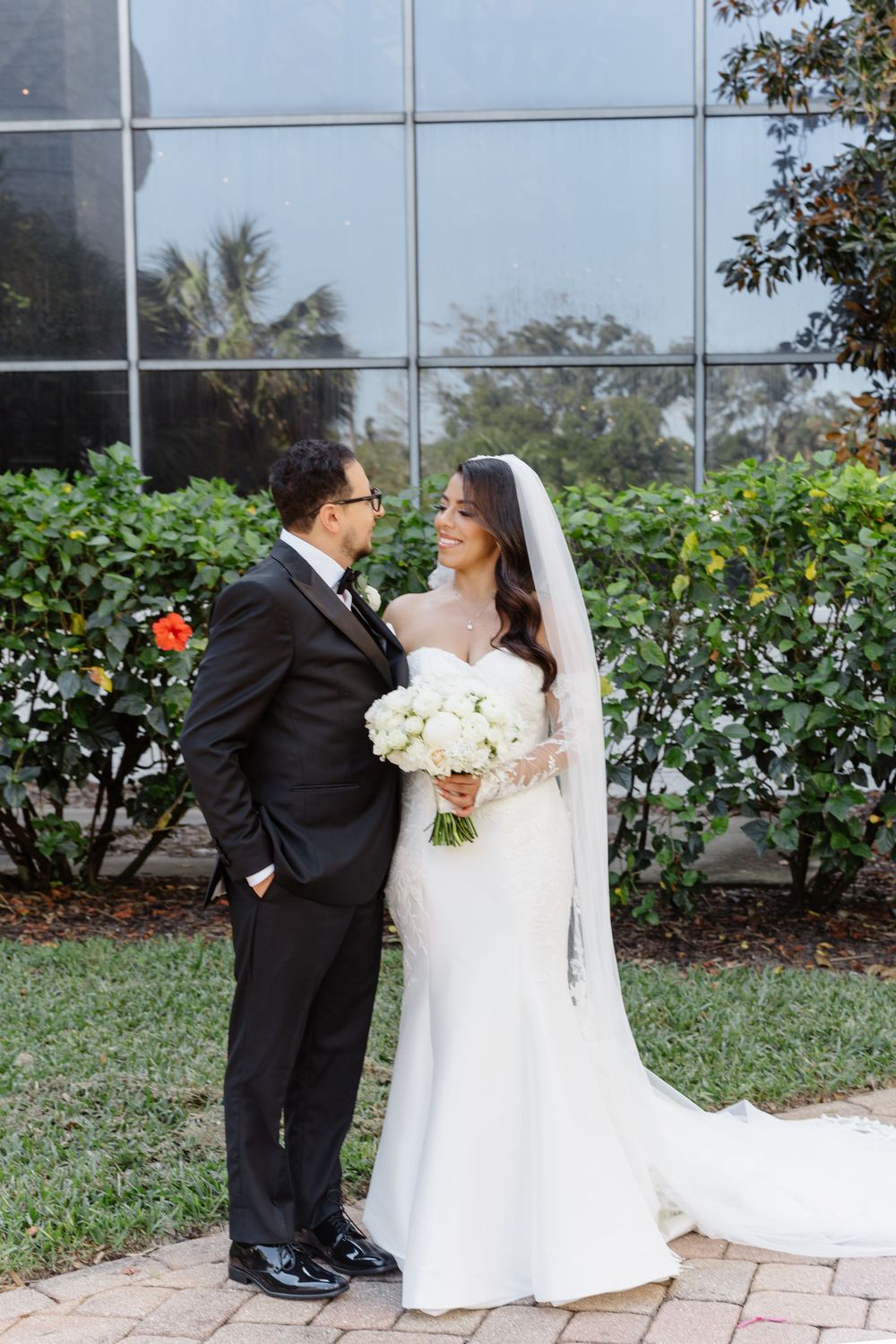 A wedding couple shares a romantic moment in their formal attire standing outside a glass building.