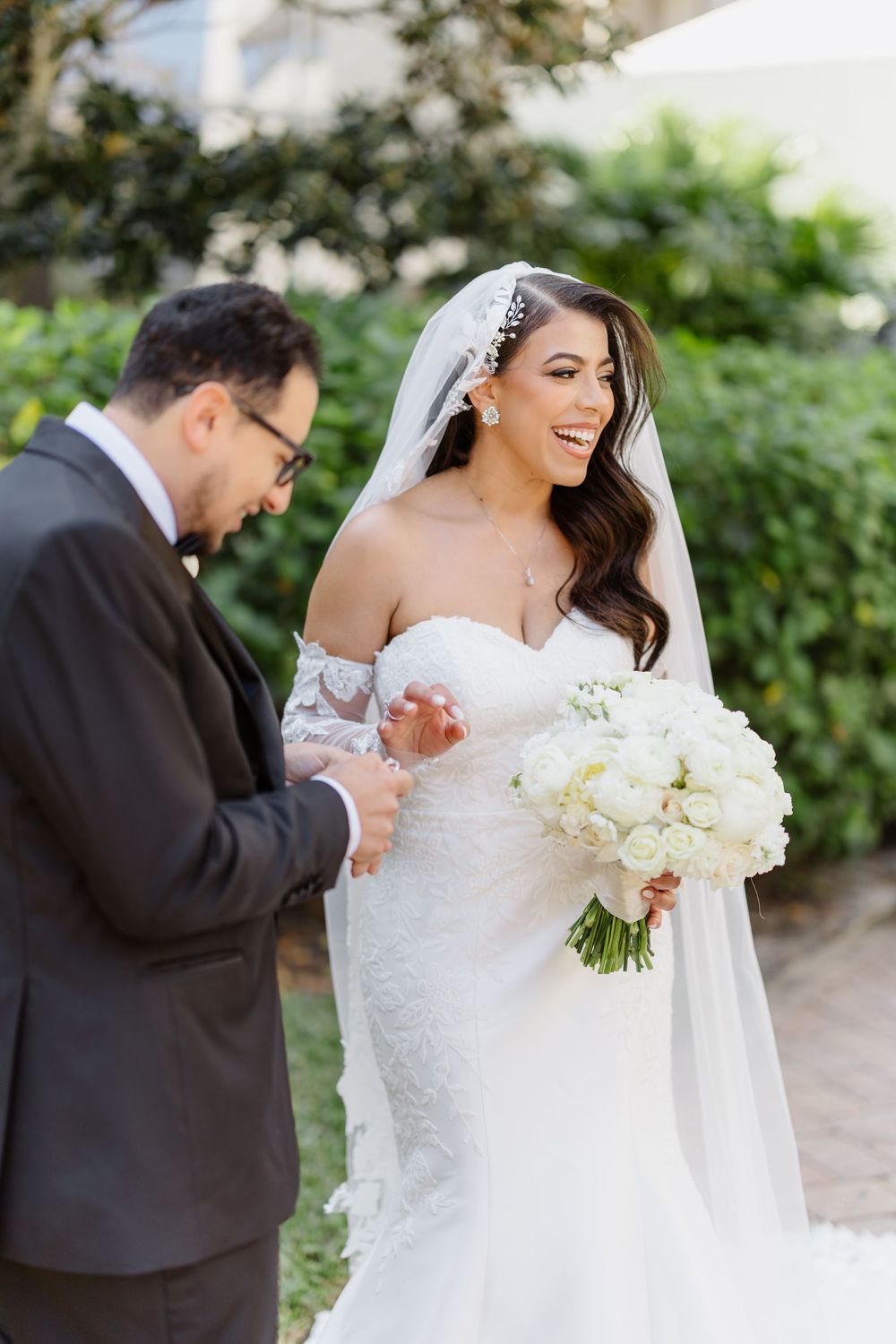 A bride in a white lace gown holds a bouquet of white flowers while sharing joyful moments in a garden setting.