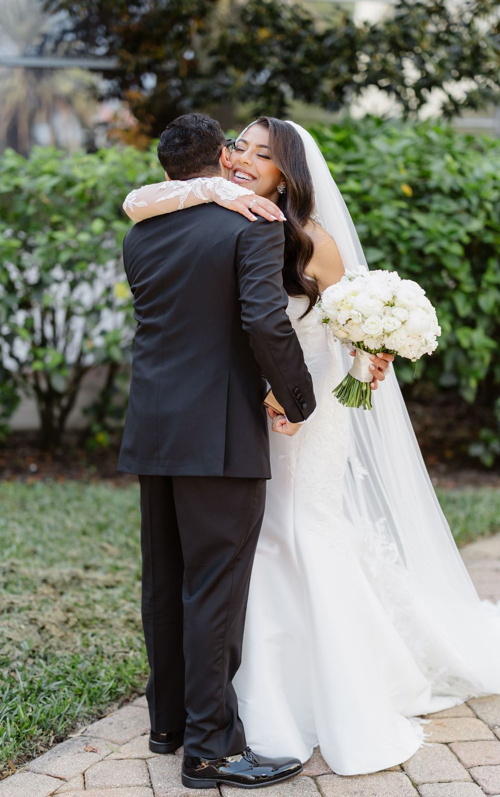A bride in a white wedding gown embraces her groom while holding a white floral bouquet during an outdoor garden ceremony.