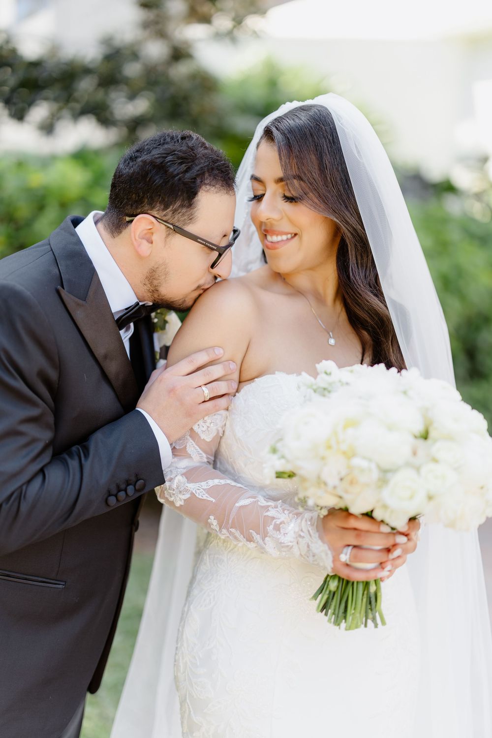 Wedding couple shares tender romantic kisses while holding a beautiful white bouquet in outdoor garden setting.