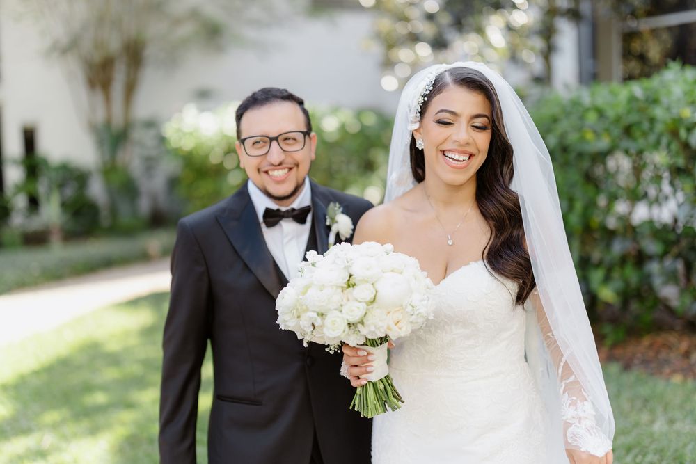 Elegant wedding couple in formal attire poses together in outdoor garden setting with white bouquet.