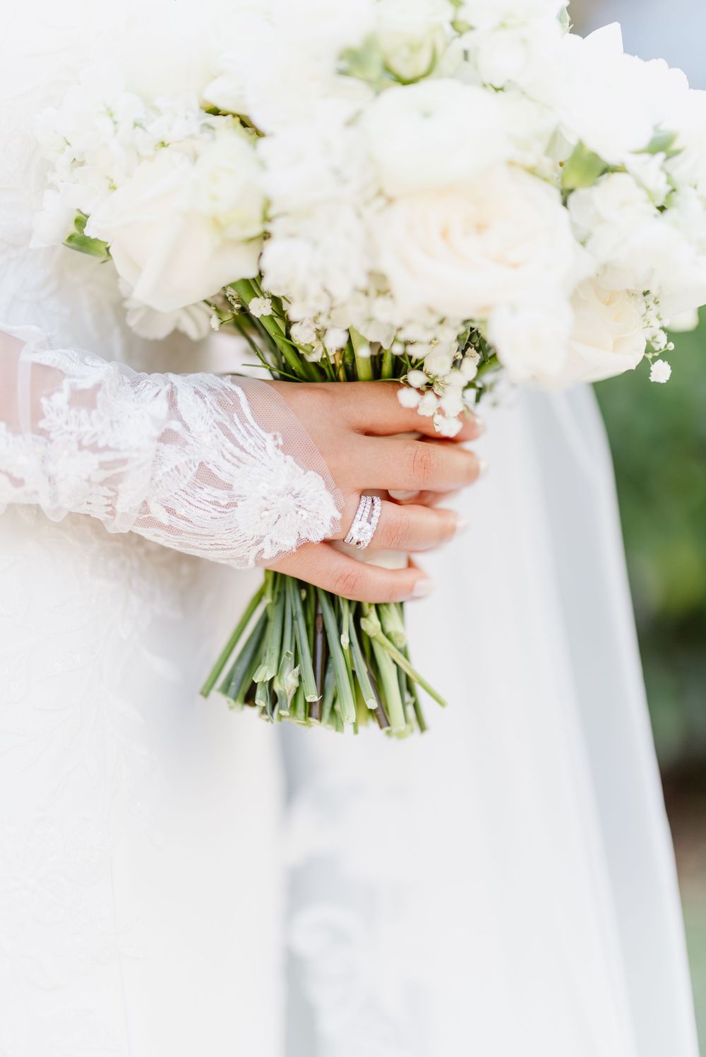 A wedding bouquet of white flowers wrapped in delicate lace is held against a flowing white dress.