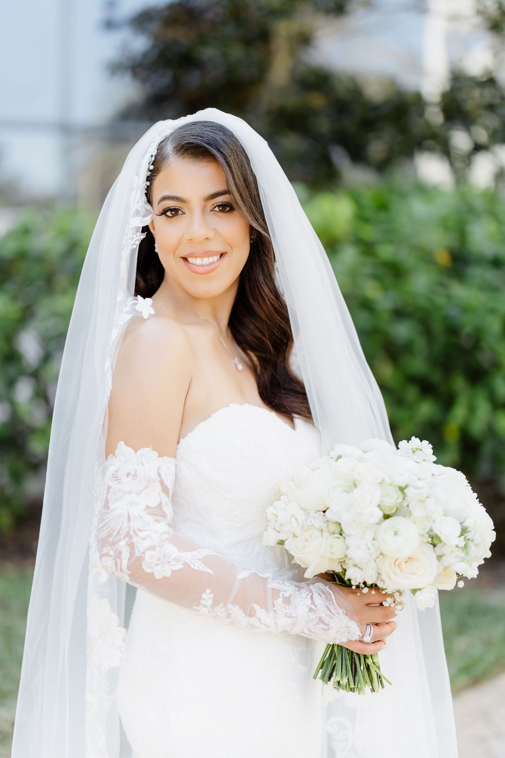 A radiant bride in a white lace wedding gown with long veil holds a beautiful bouquet of white flowers in outdoor portraits.