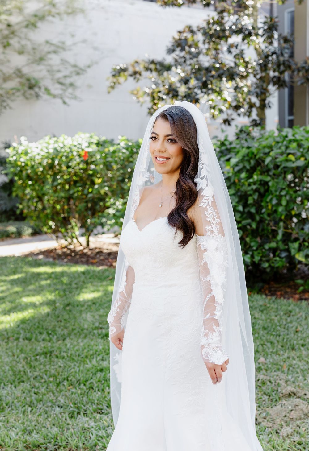 Bride in a romantic long sleeve lace wedding gown poses on a sunny day against a garden backdrop.
