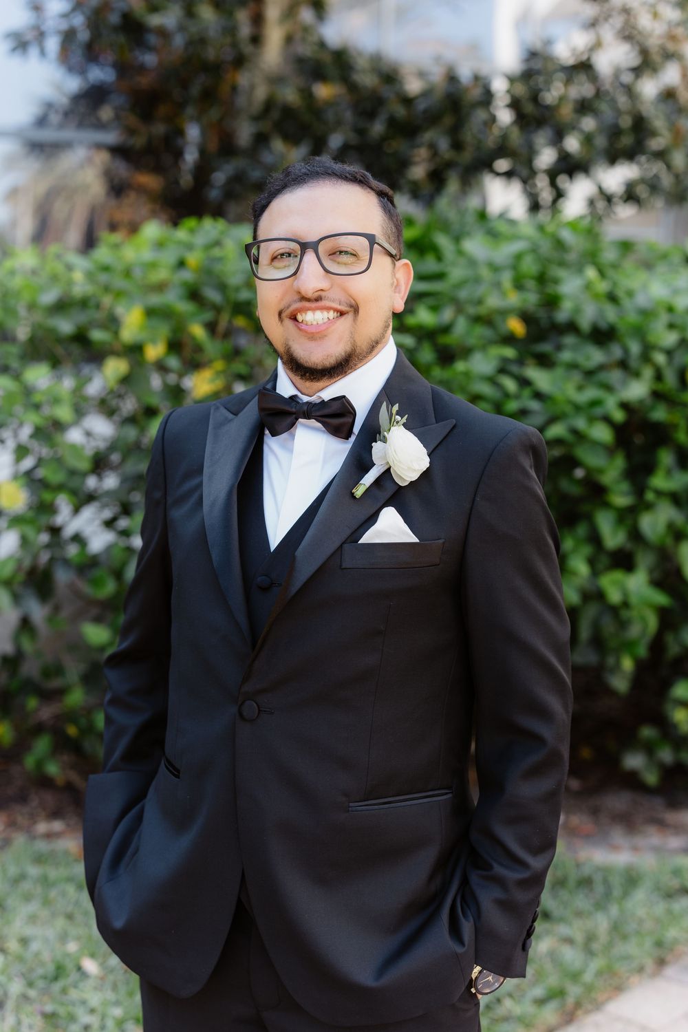 An elegantly dressed person in a black tuxedo with bowtie and glasses smiles while standing in a garden setting.