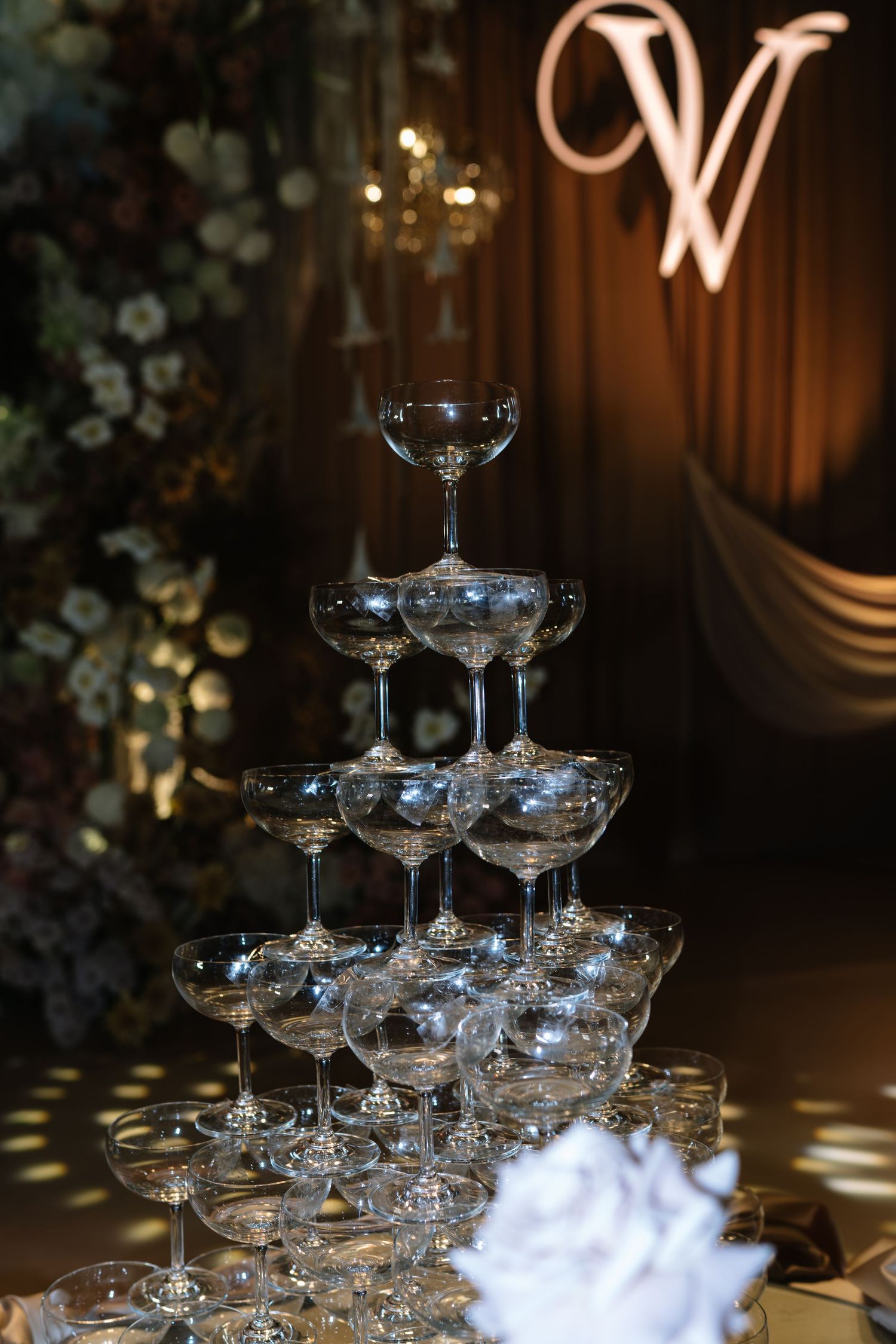 Pyramid tower of champagne coupe glasses at a wedding reception with floral and monogram backdrop.