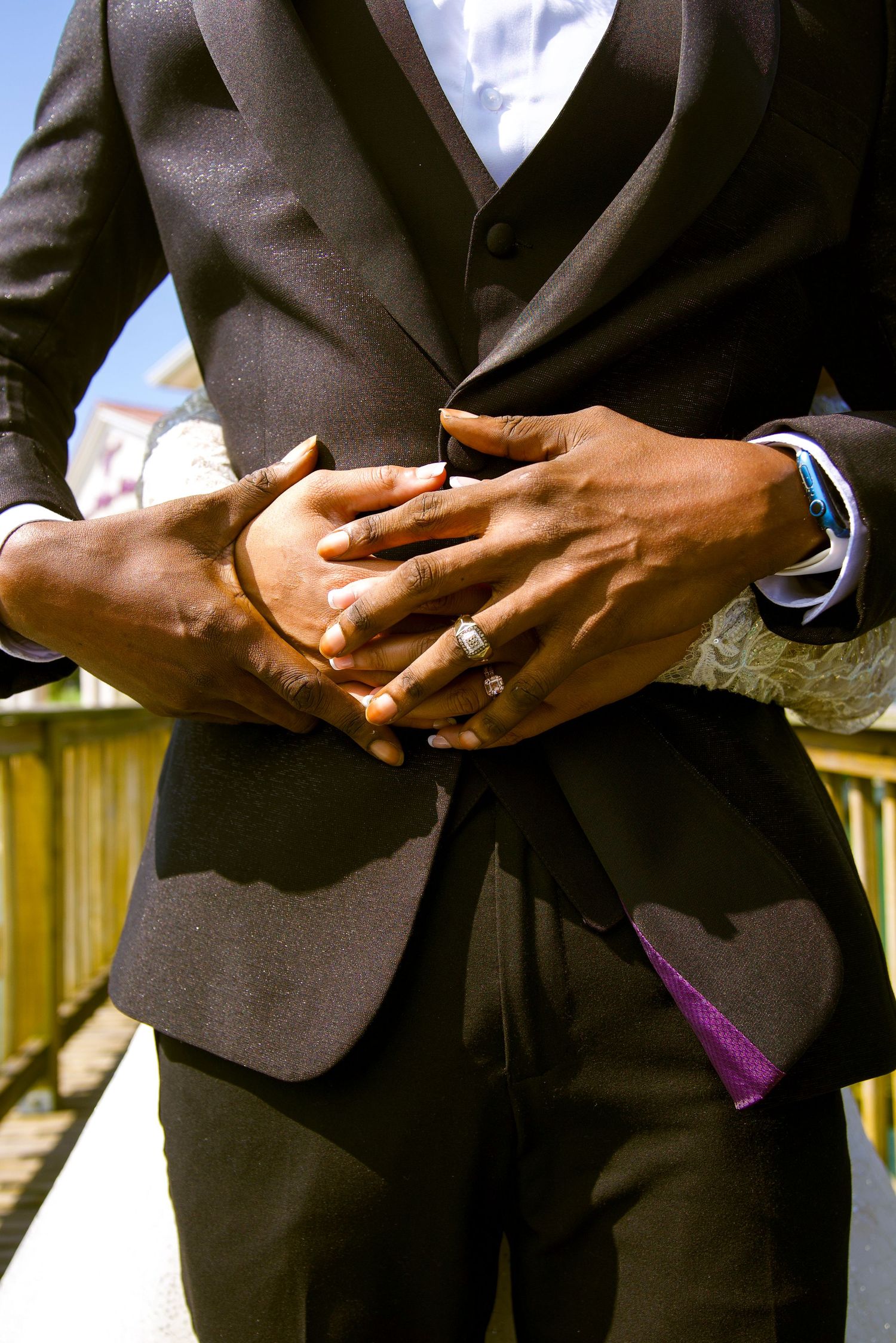 Close-up of hands clasped over a black suit jacket showing wedding rings and formal attire.