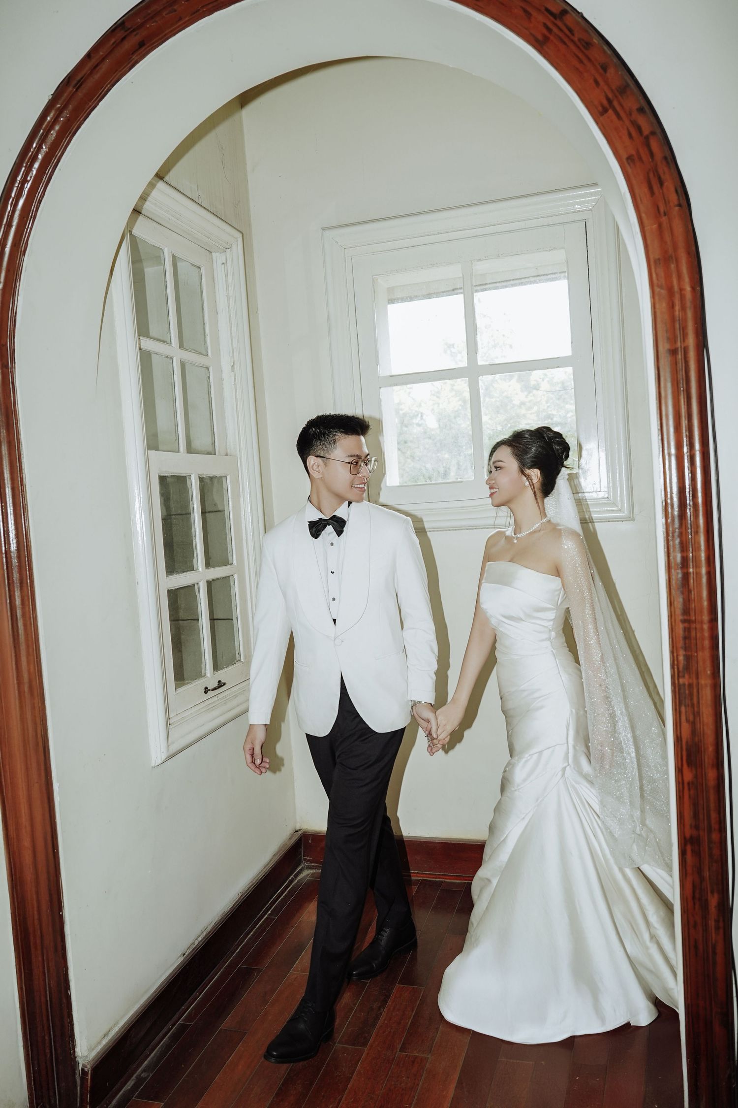 Bride and groom hold hands walking through an elegant arched doorway in a sunlit white interior.