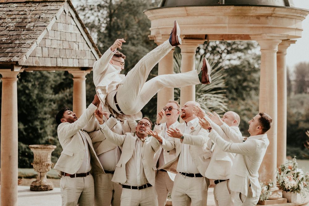 Groomsmen with tan suits raise their arms in celebration at an outdoor wedding venue with white columns.