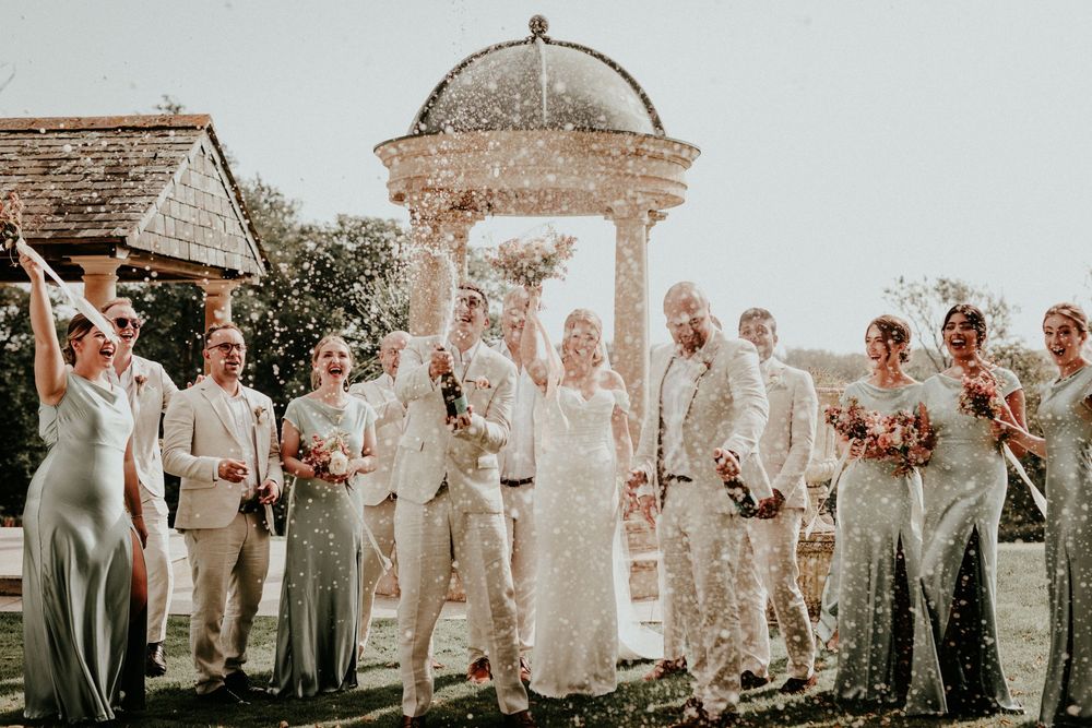 Wedding party members wearing sage green and ivory dresses pose outdoors in an elegant garden setting on sunny day.