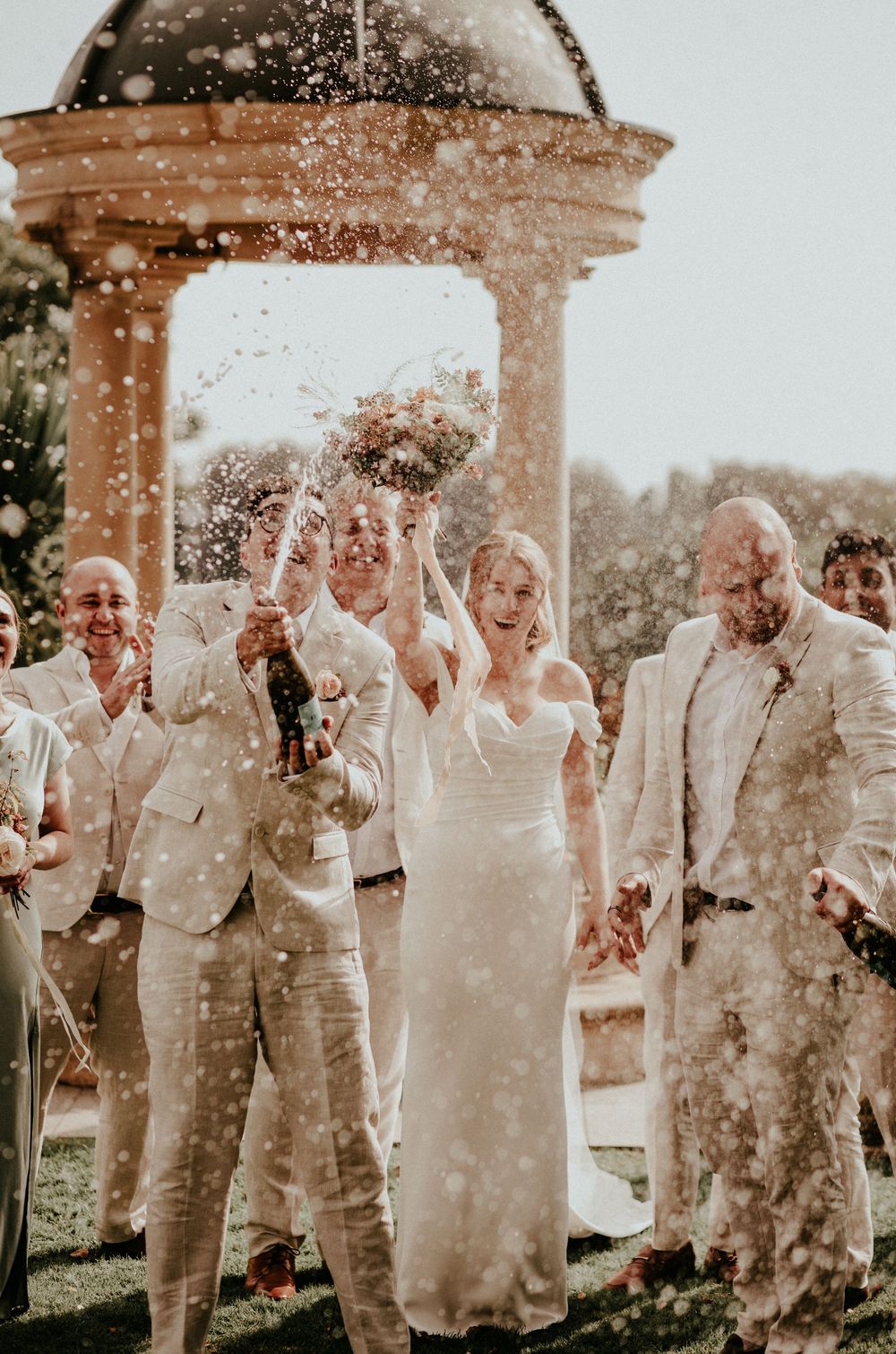 A joyous celebration under a stone gazebo as confetti showers down on wedding guests in light-colored attire.