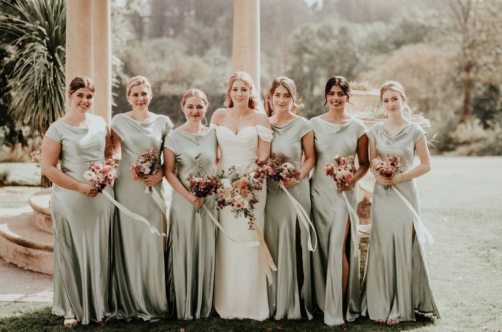 Wedding party in sage green dresses poses outdoors on a garden lawn for a group photo.