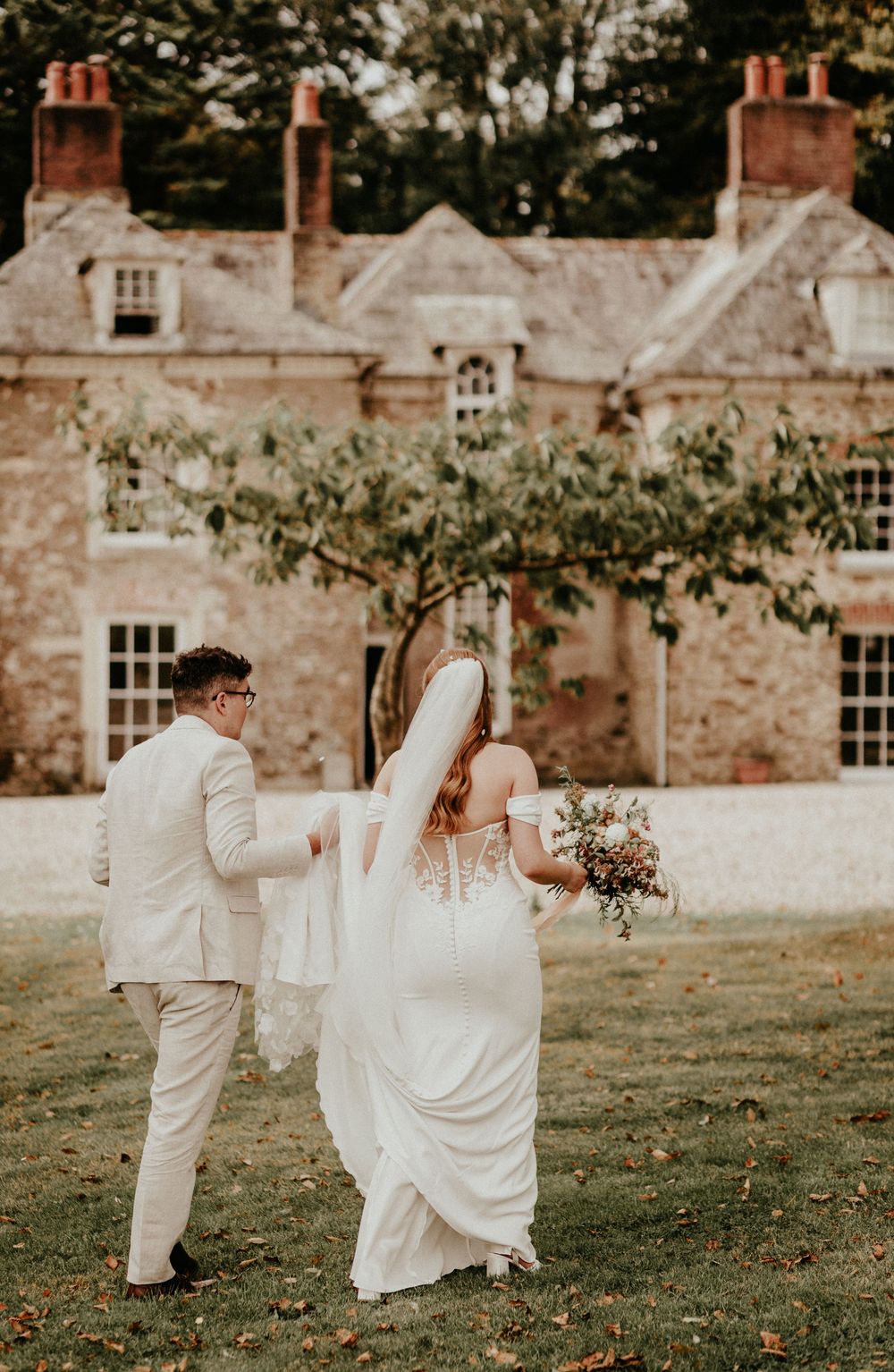 A couple holds hands while frolicking across a grassy lawn at a rustic stone manor wedding venue.