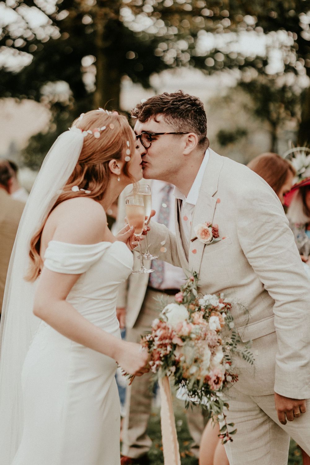A romantic outdoor wedding moment at sunset as couple shares kiss while bride holds delicate floral bouquet.