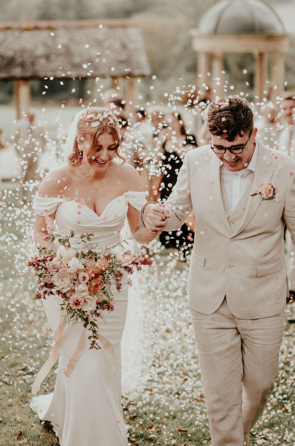 Newlywed couple wearing light beige and white attire walk together in a dreamy garden setting with soft lighting.