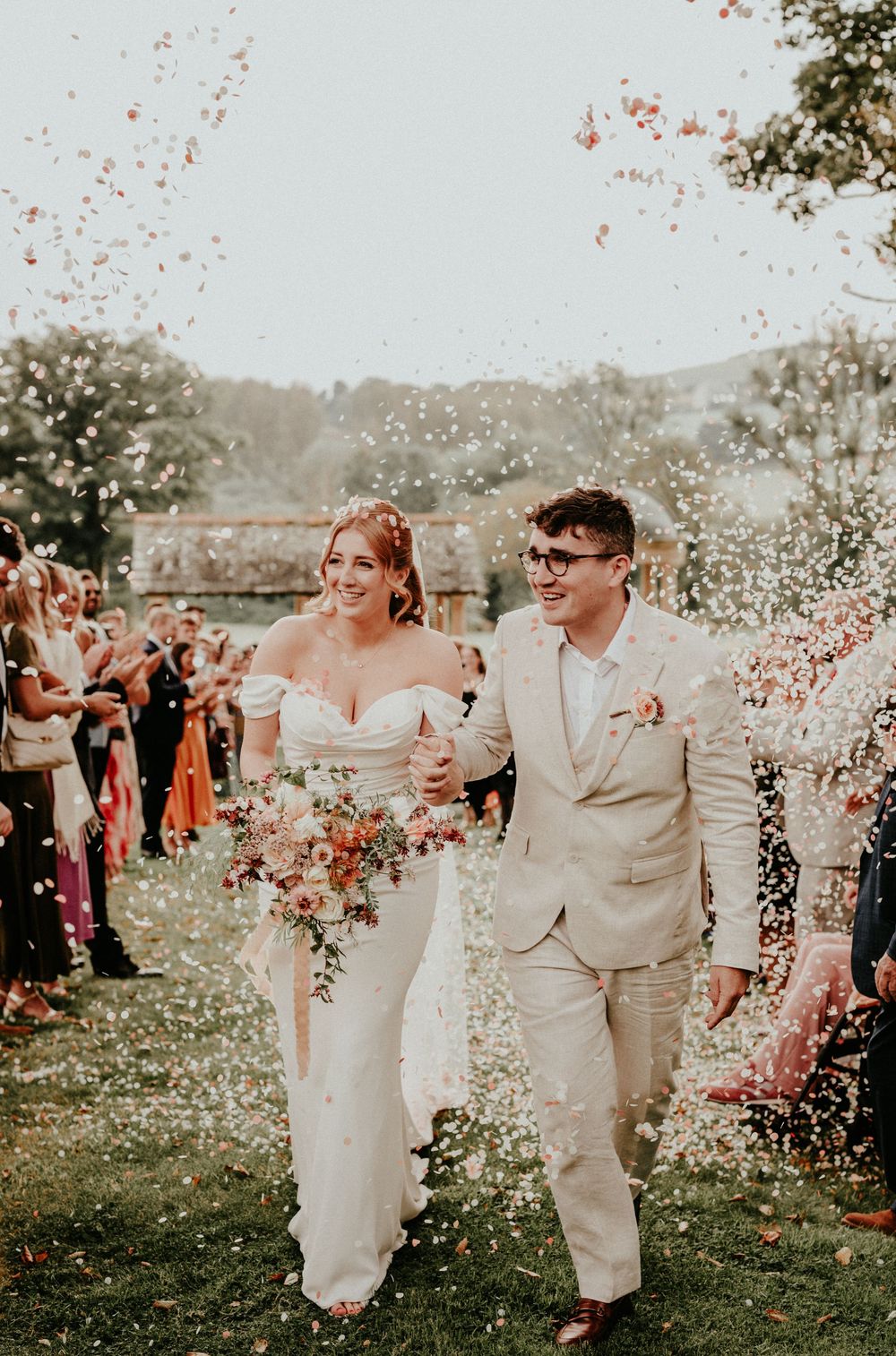 A couple celebrates during their outdoor wedding ceremony as flower petals shower from above on a sunny day.