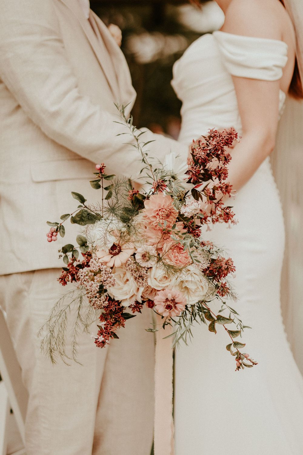 Wedding party in beige attire holds elegant pink and burgundy floral bouquets against neutral background.