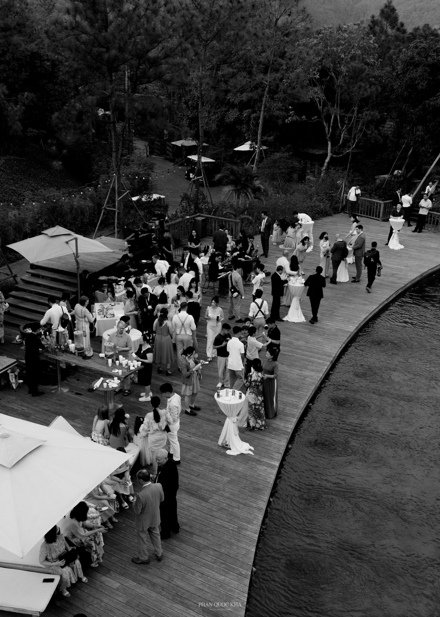 Aerial black and white photo of wedding cocktail hour guests on a wooden deck surrounded by lush trees.