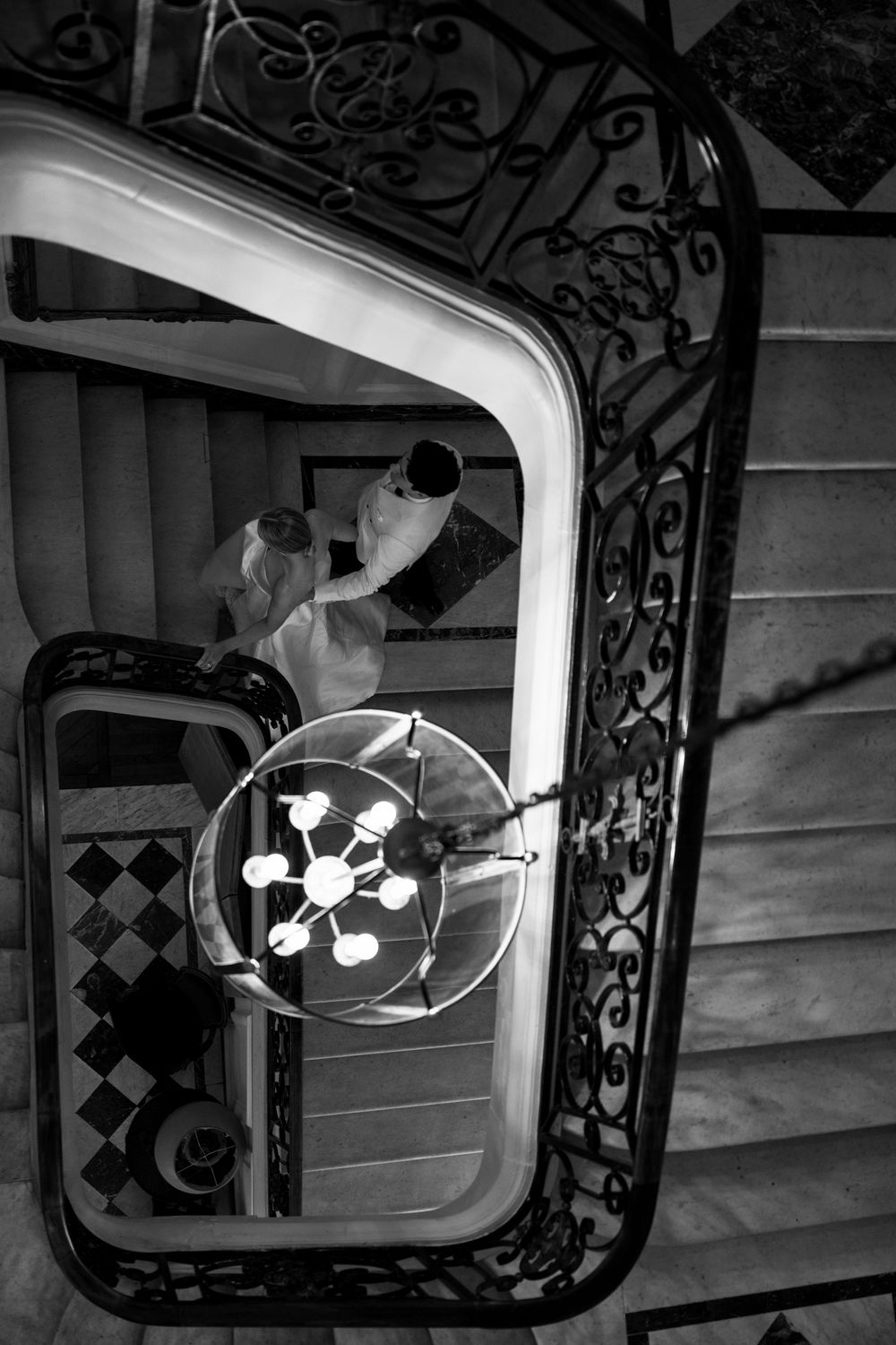 A black and white image shows an ornate spiral staircase with decorative metal railings and overhead lighting fixtures.