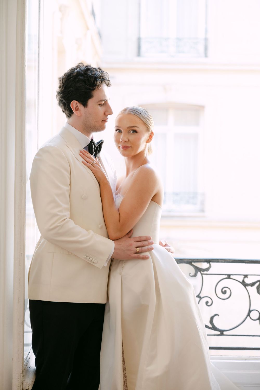 A romantic couple in white attire share intimate moments by a window in a bright, airy space.