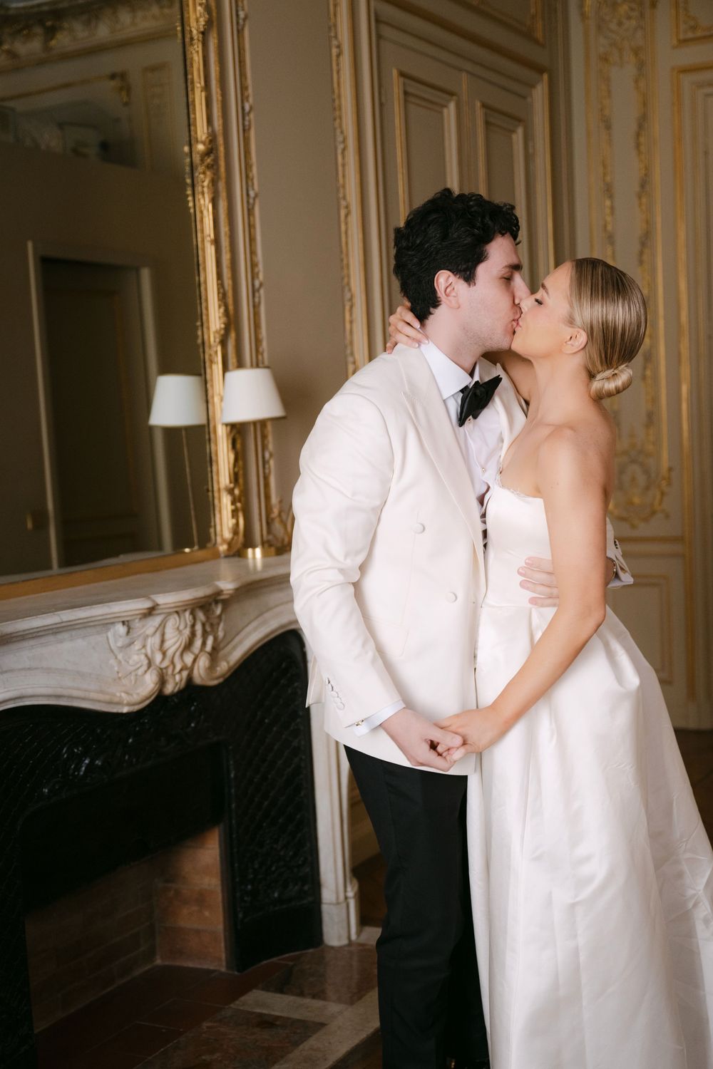 A romantic wedding couple shares an intimate moment in an elegant room with ornate gold mirrors and a fireplace.