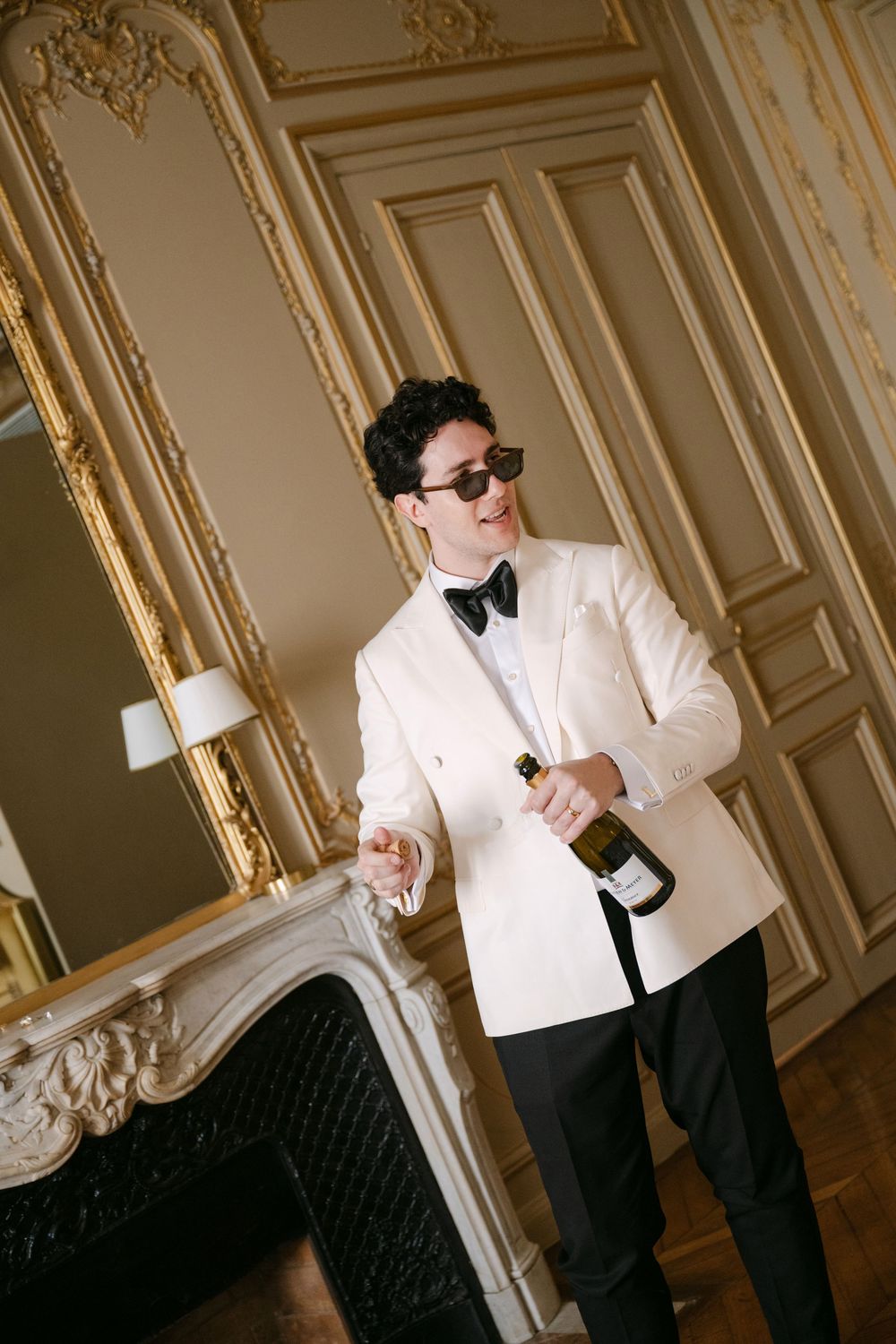A person in a white dinner jacket and bow tie holds a champagne bottle in an ornate room with gilded wall panels and a fireplace.