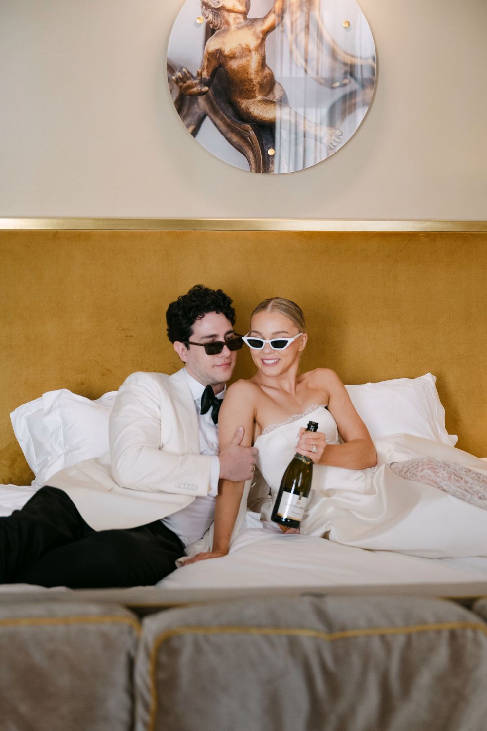 A couple in formal attire lounges together on a bed with champagne and white linens.