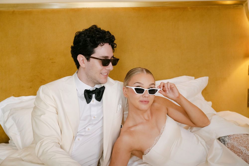 A wedding couple in white attire laughs together during a candid moment with a warm golden background.