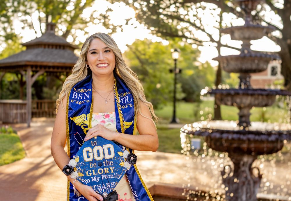 Smiling graduate in blue and gold BSN sash poses outdoors near fountain at golden hour.