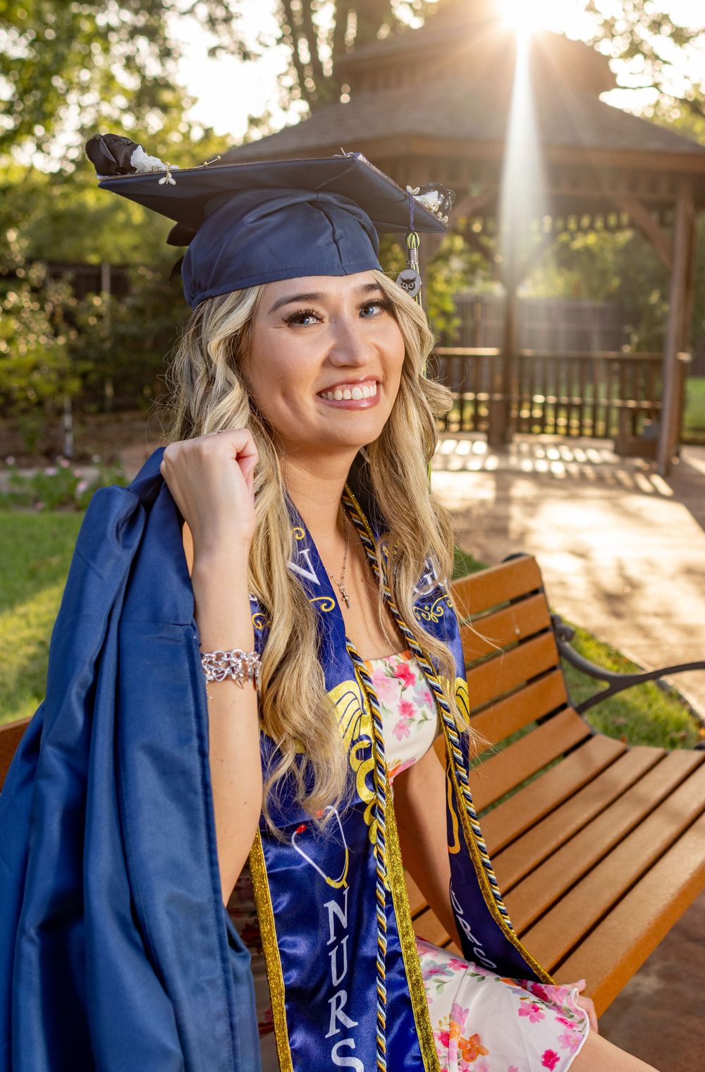 Smiling nursing graduate in blue cap and gown with gold cords sits on bench outdoors in golden sunlight.