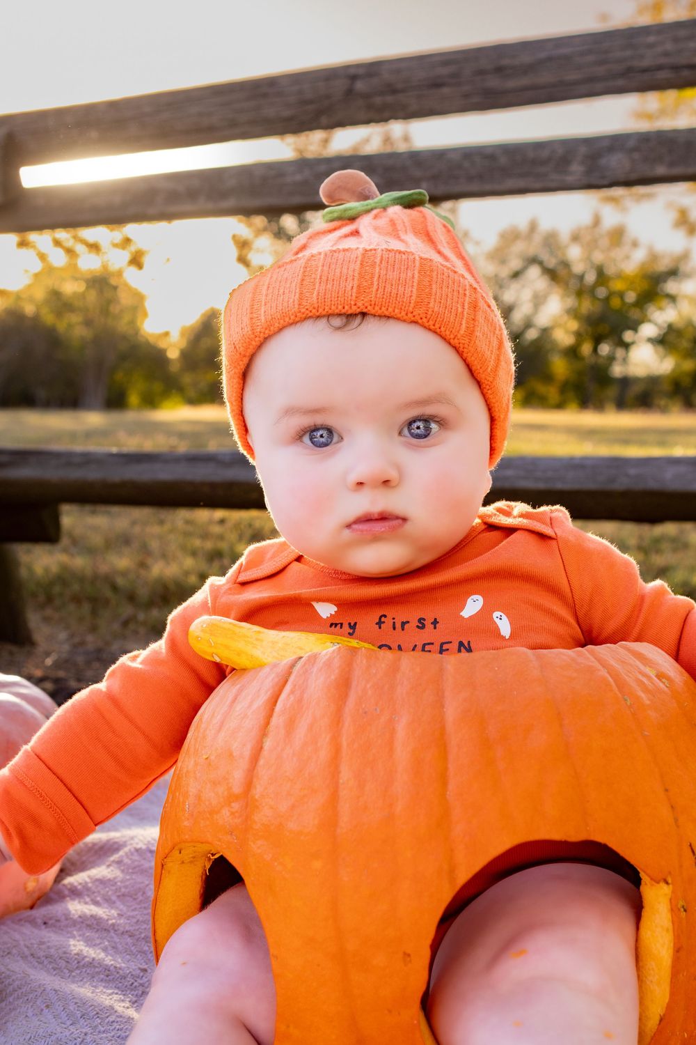 A smiling baby in an orange pumpkin outfit posed with a real pumpkin outdoors.