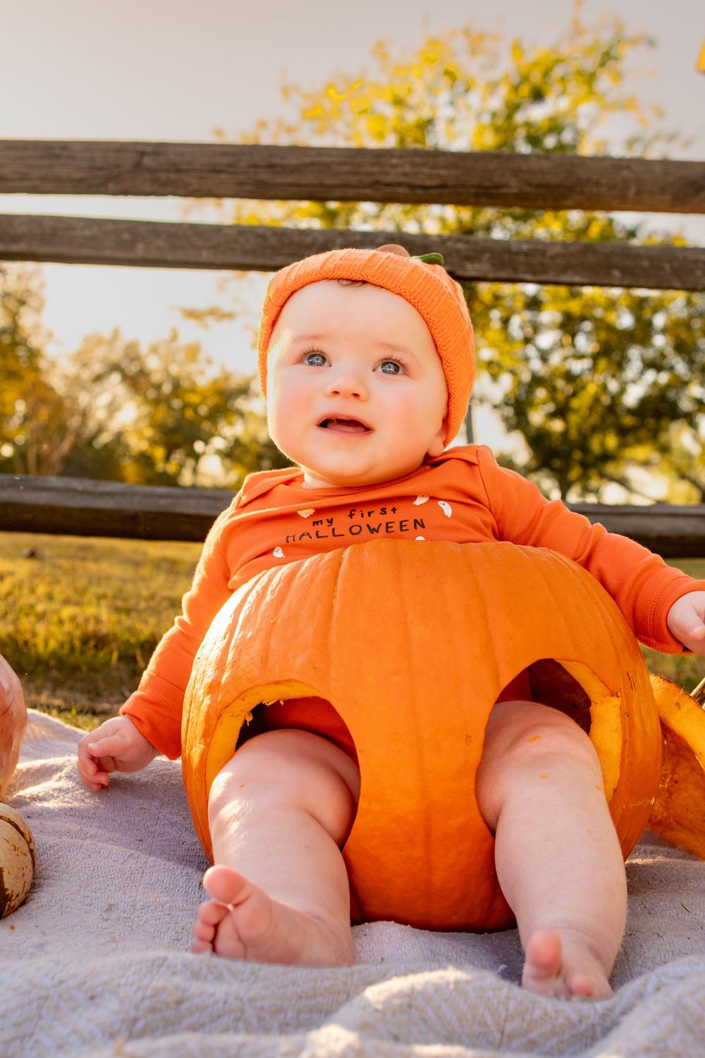 A baby wearing an orange pumpkin costume lying on a blanket outdoors.