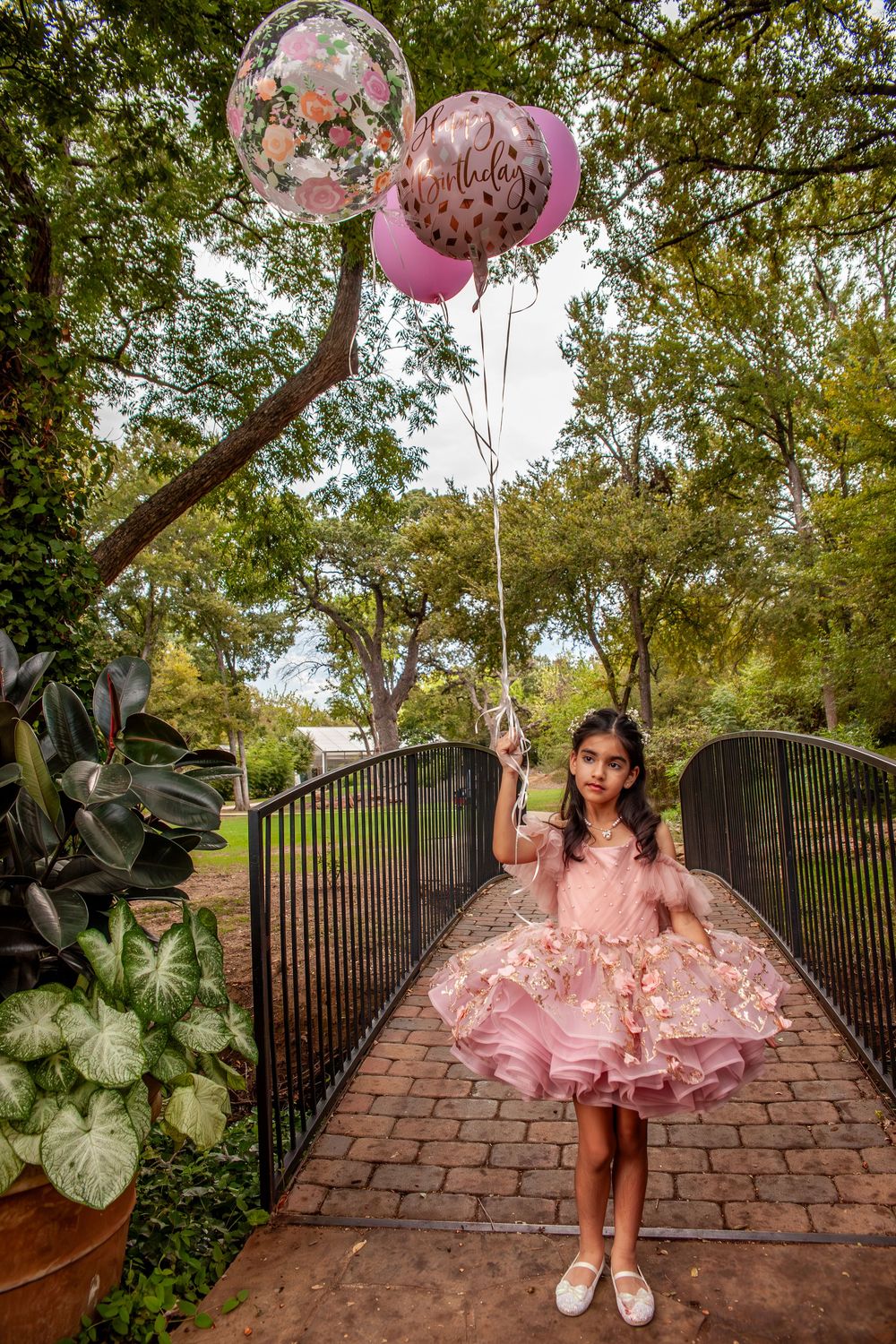 Pink tulle dress twirls on brick pathway surrounded by lush garden and balloons.