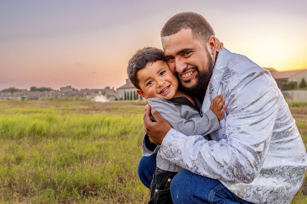 A father and young son share a warm hug outdoors at sunset, smiling joyfully in a green field.