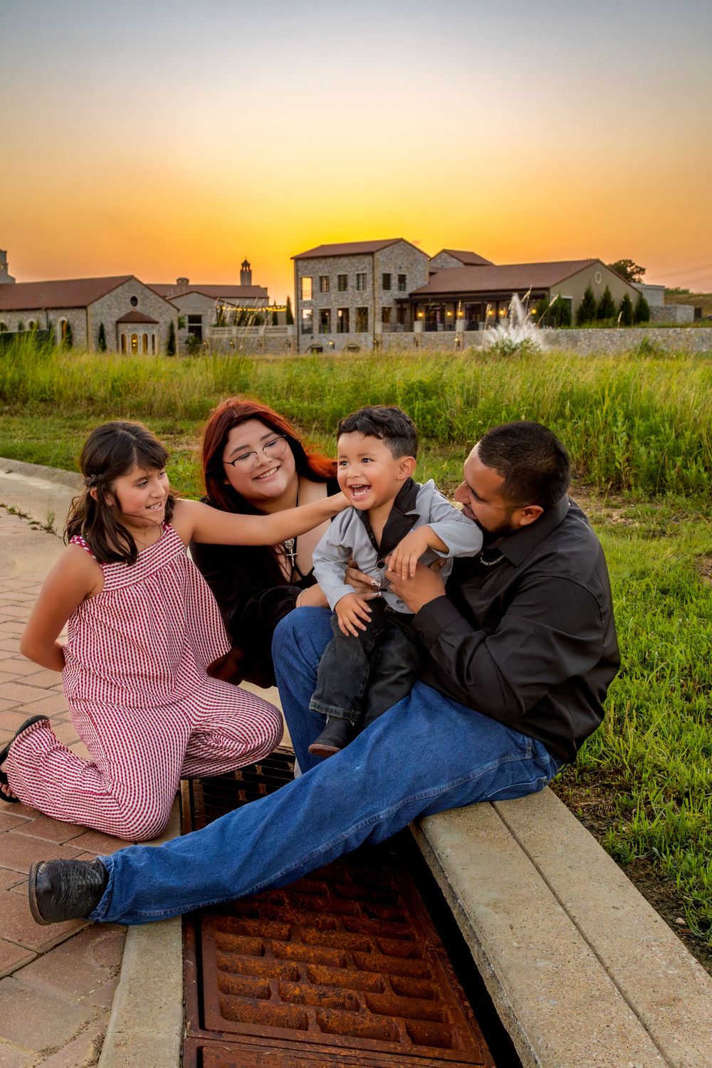Family of four laughing together at sunset near a fountain and large estate building in a warm evening glow.