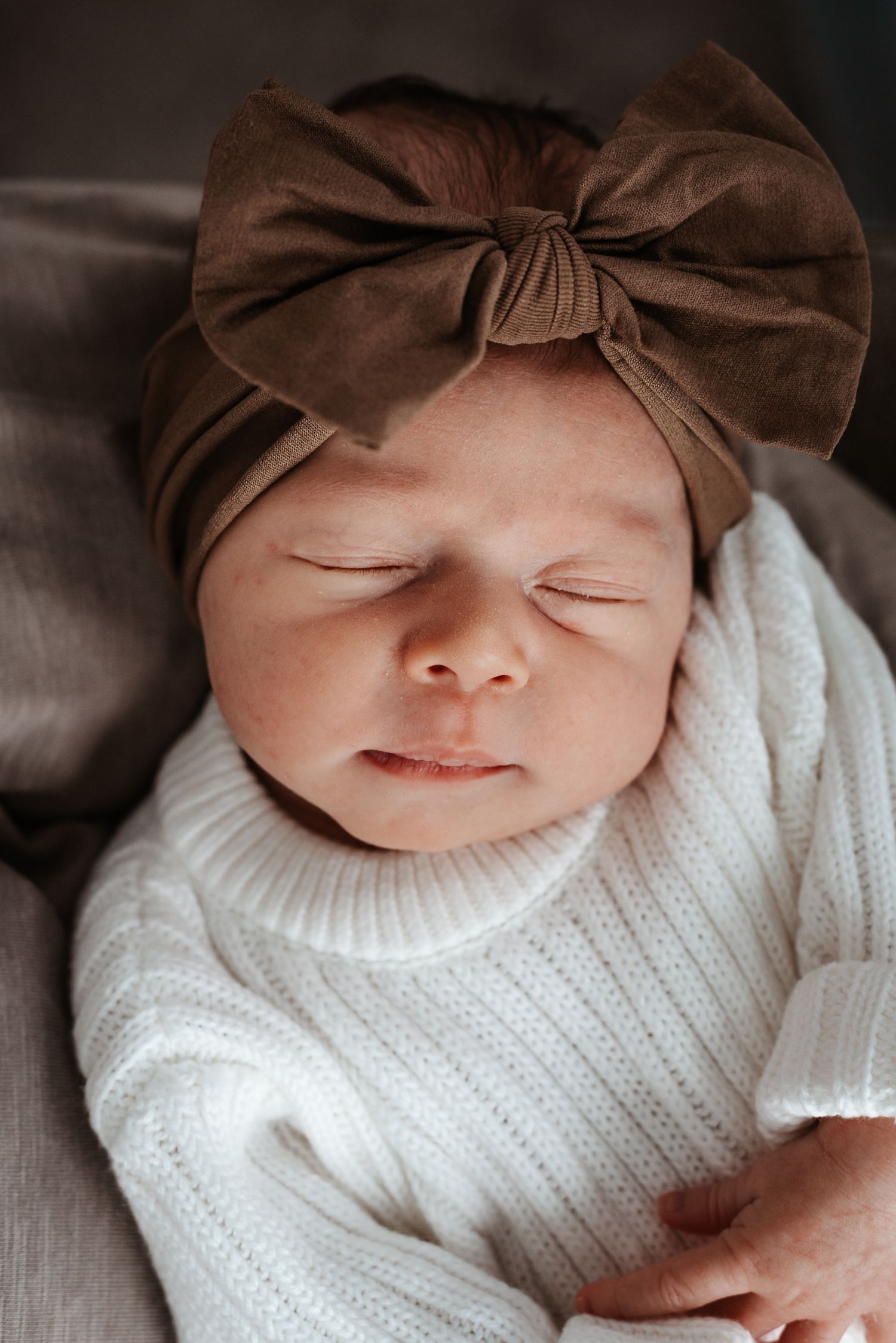 Sleeping infant wearing white knit sweater and brown bow headband lying on gray cushion.