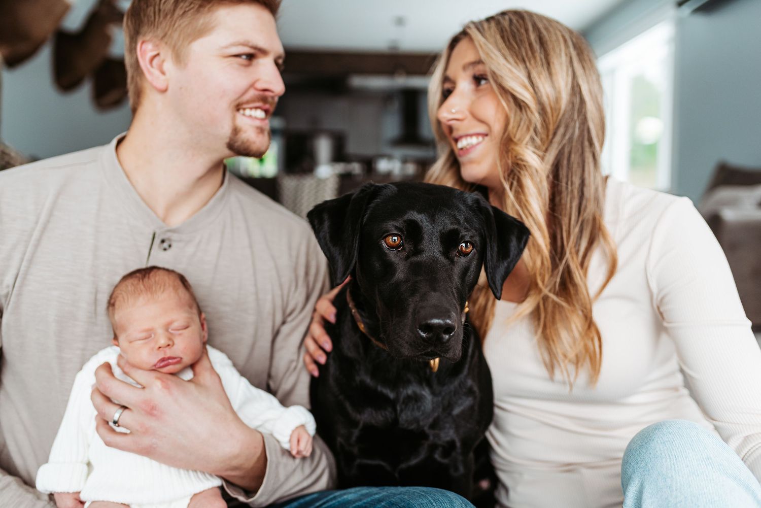 A couple sitting on couch with their newborn baby and black Labrador dog sharing a happy family moment.