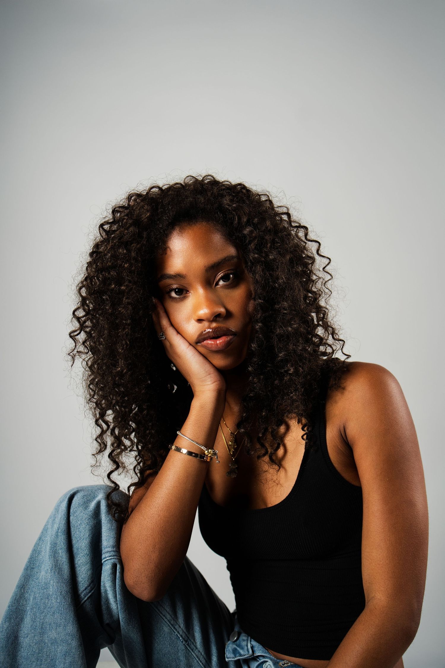 Portrait of a person with natural curly hair wearing a black tank top and denim, resting their face on their hand against gray background.