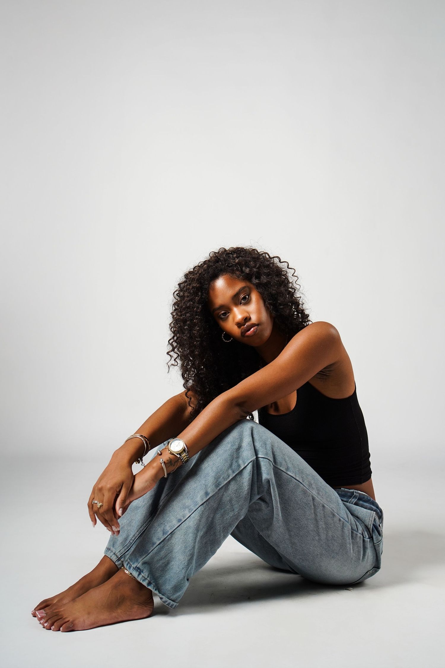 A person in a black top and light jeans sits curled up in a contemplative pose against a white studio backdrop.