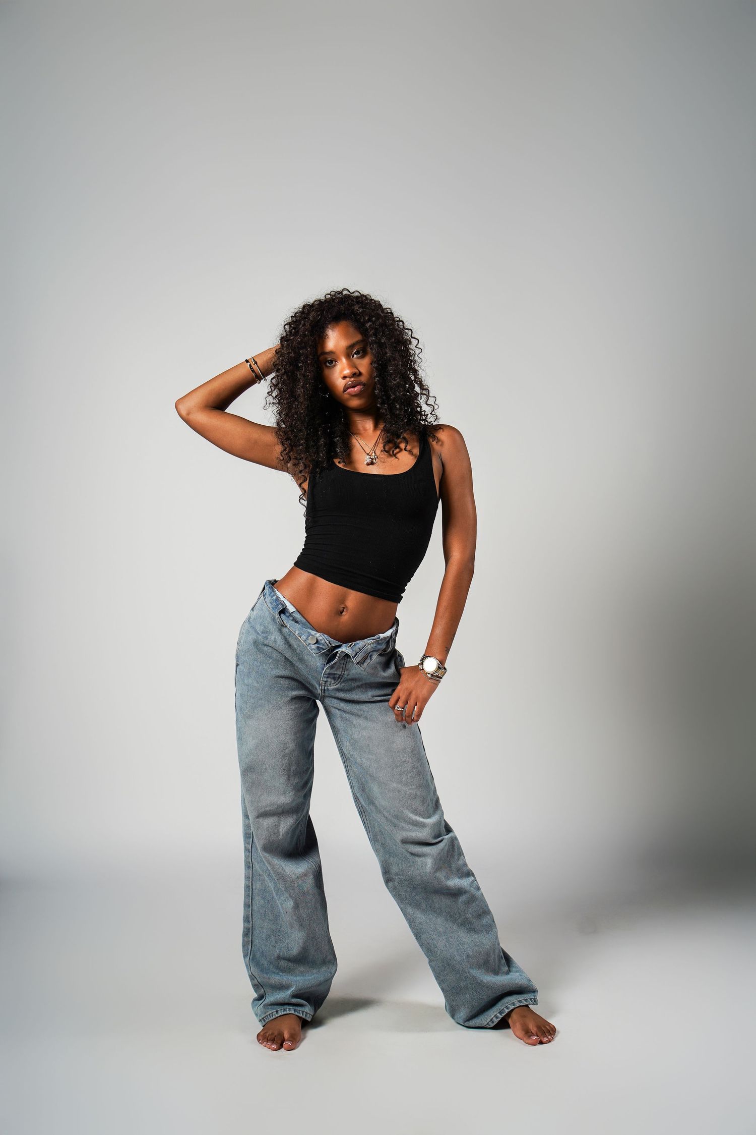Fashion model poses dynamically in baggy blue jeans and black crop top against white studio background.