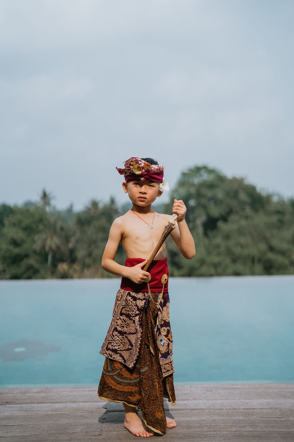 A child in traditional Balinese attire poses on a wooden deck overlooking turquoise waters and tropical landscape.