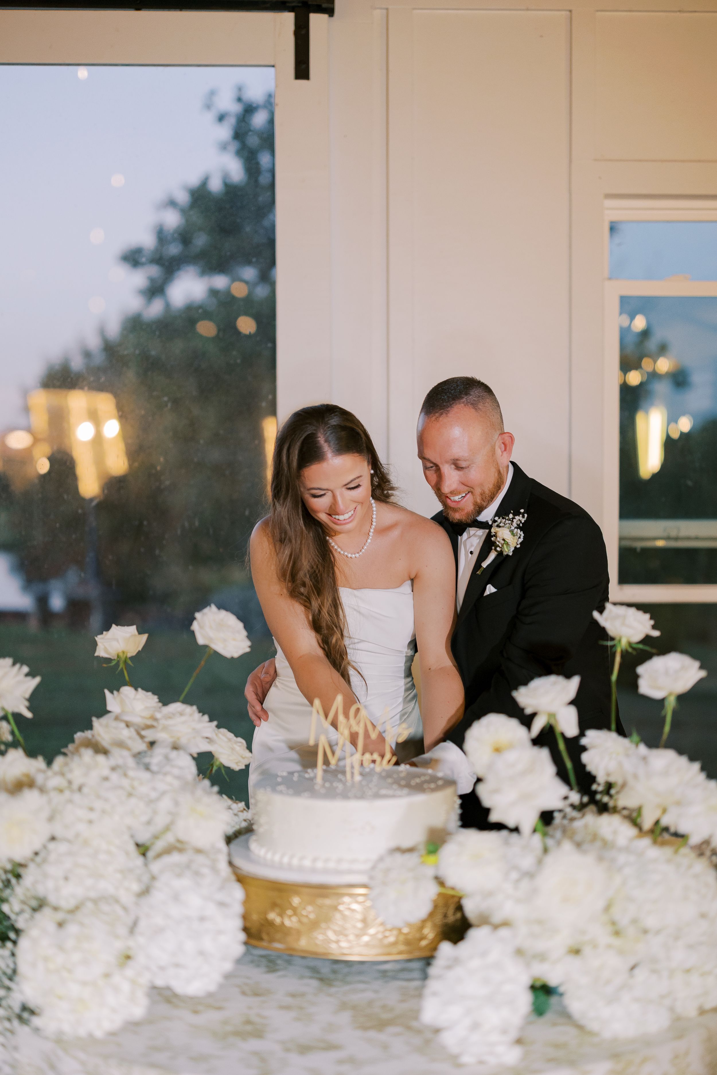 Wedding couple cuts their cake surrounded by elegant white floral arrangements at an indoor reception venue.