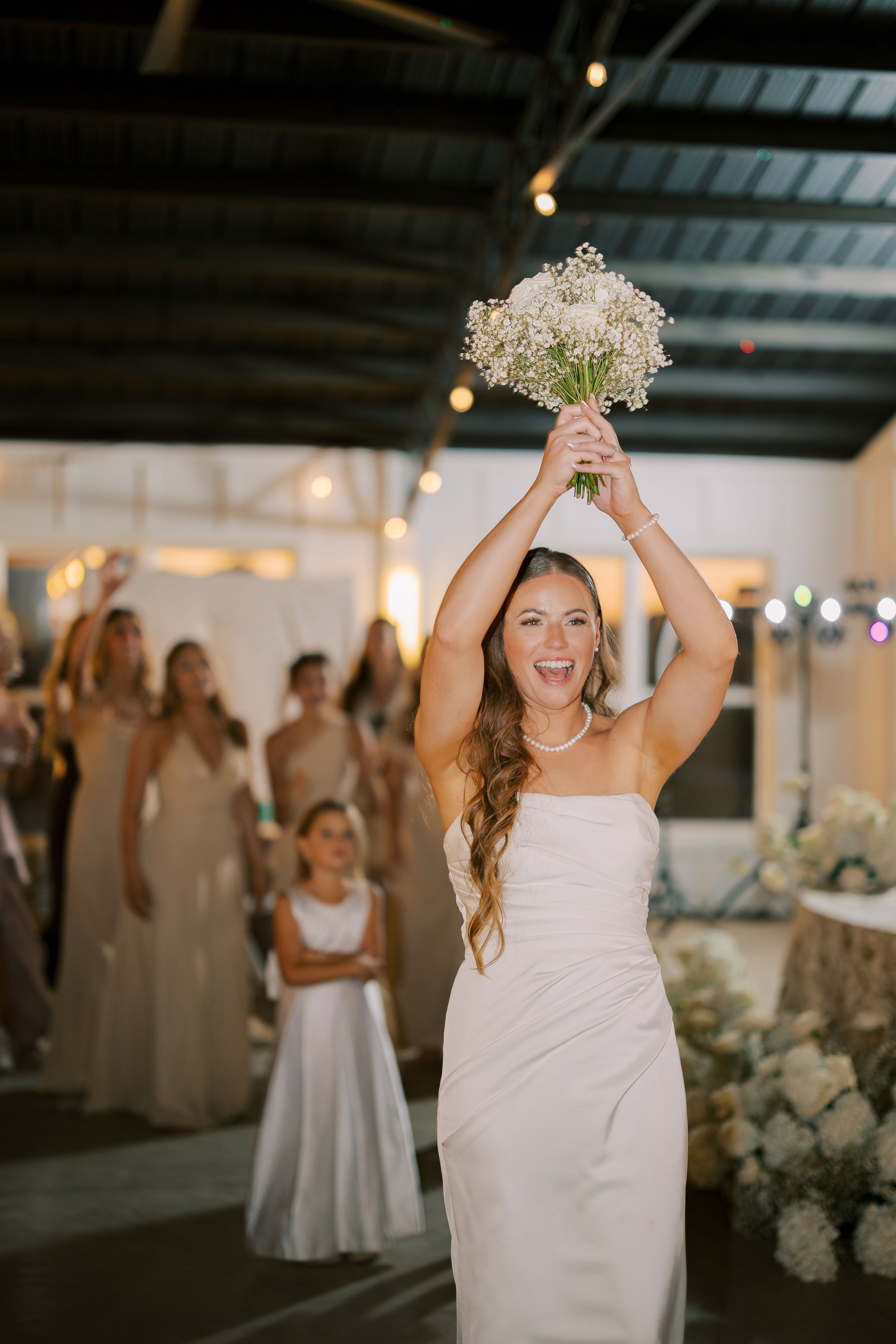 Wedding party celebrates in a bright indoor venue decorated with white florals and elegant lighting.