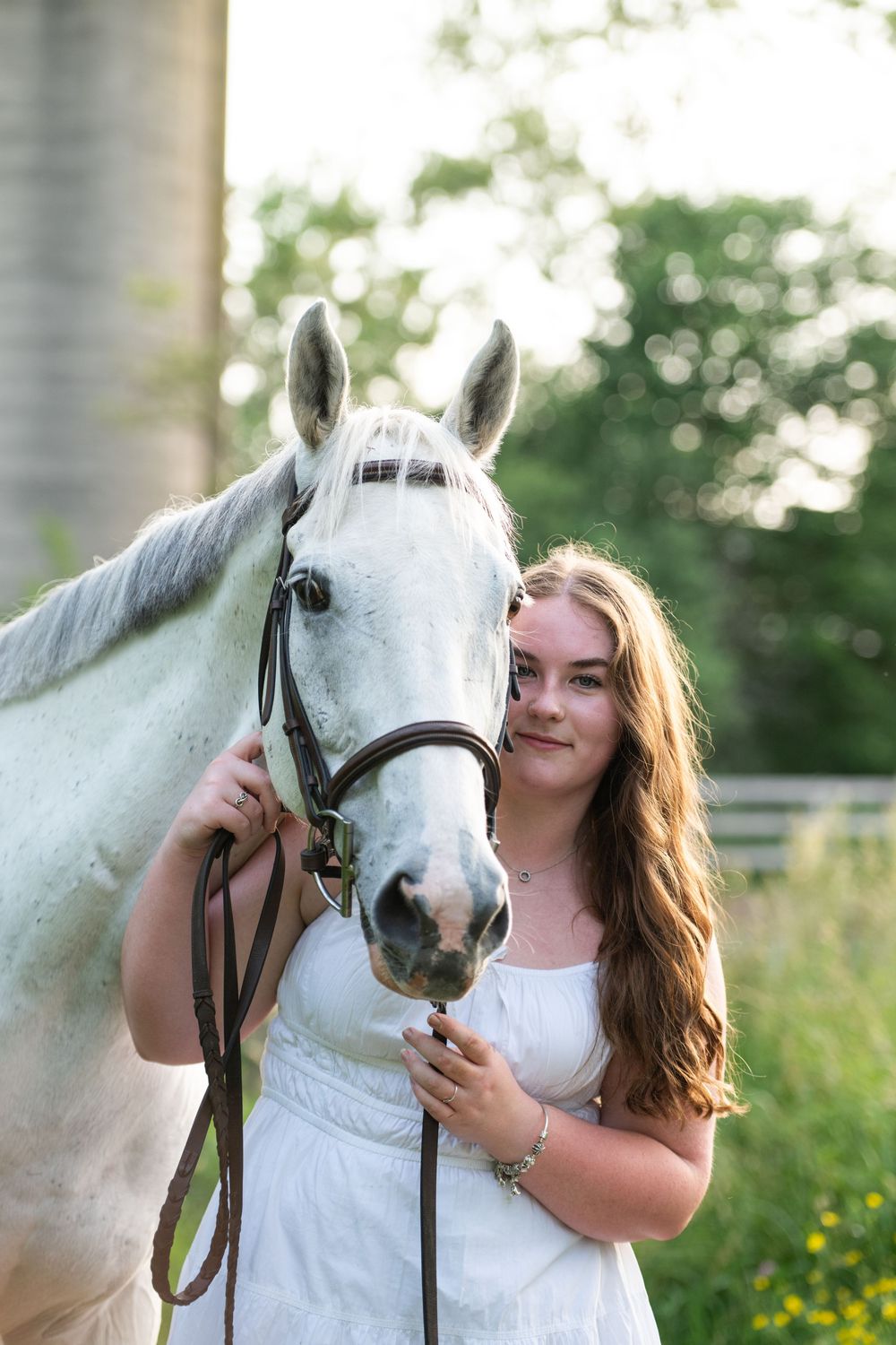 A woman in a white dress stands lovingly with her majestic white horse in a sunny meadow with wildflowers.