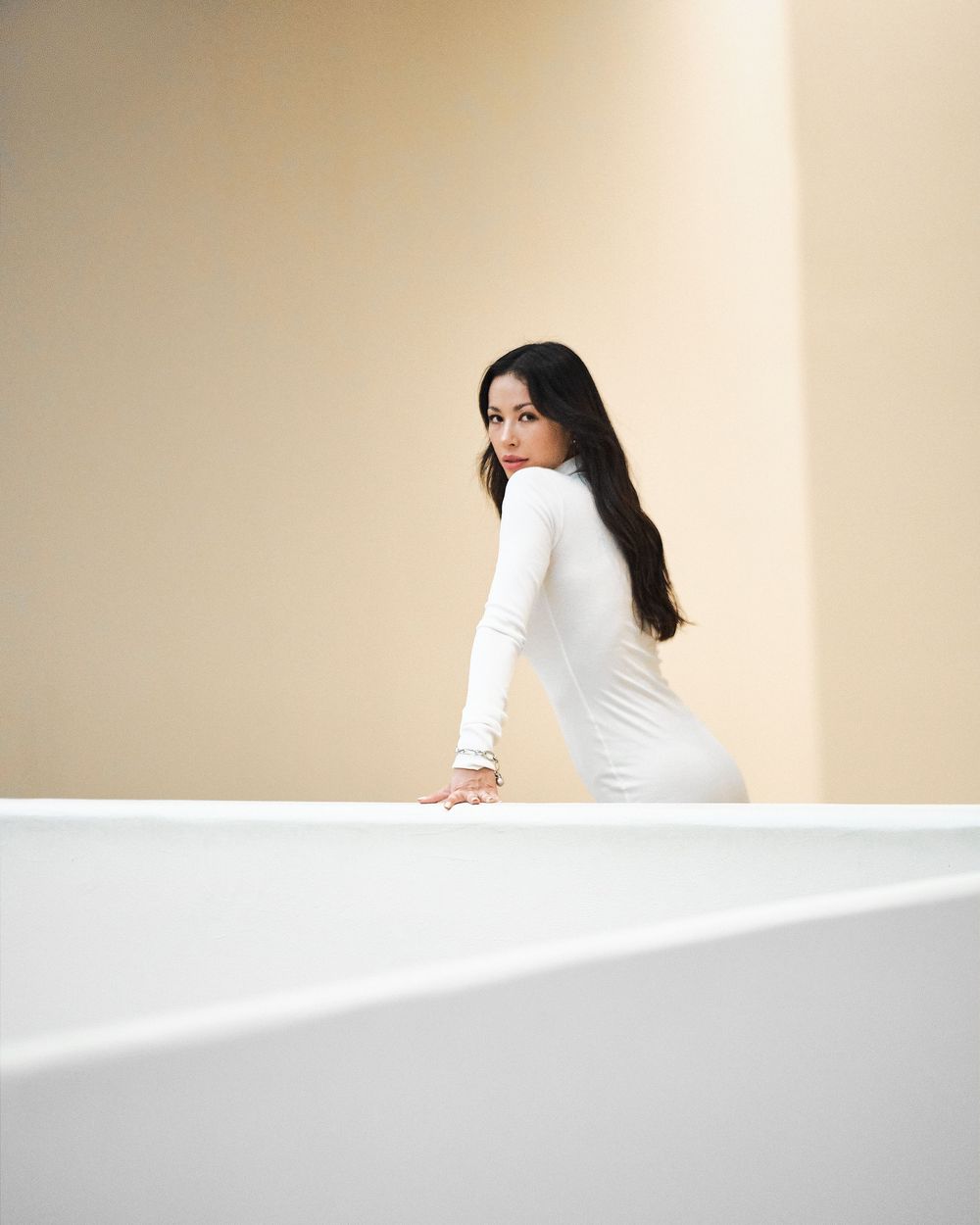 A woman in white sits against a cream wall as sunlight creates dramatic angular shadows and geometric shapes.