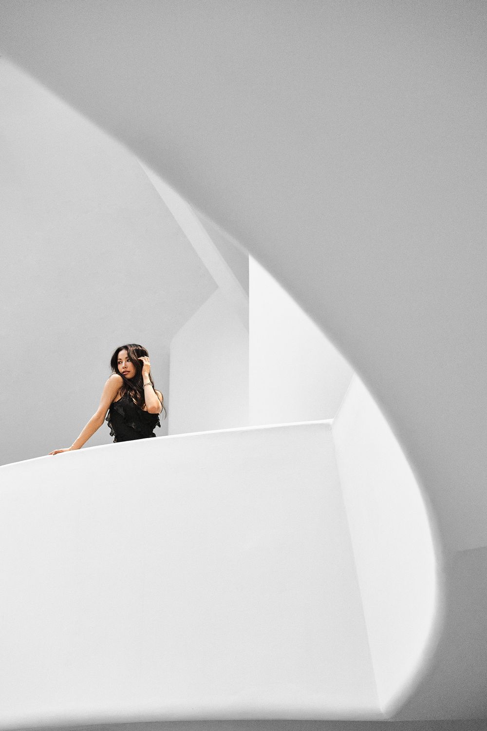 A person in black attire rests against a white curved architectural wall with dramatic natural lighting and shadows.